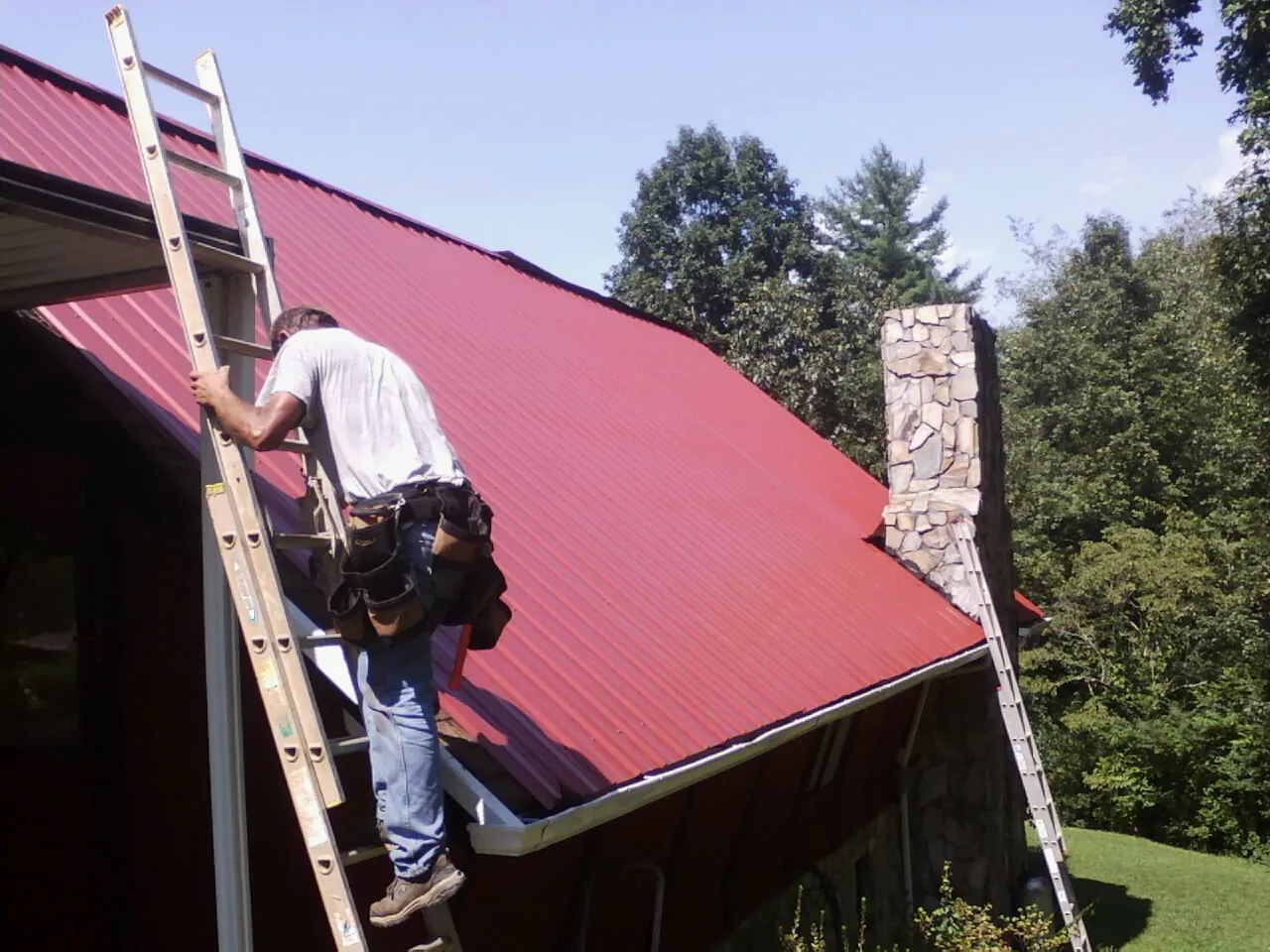 Roofer on a ladder beside a red metal roof and stone chimney — Soffit & Fascia Repair by Roofing Brothers in West Berlin